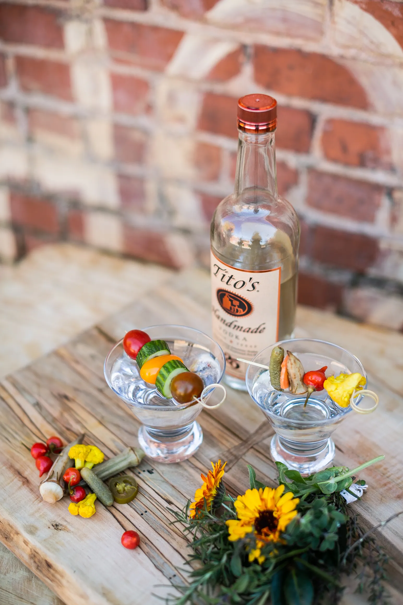 Bottle of Tito's vodka and two handmade craft cocktails with fresh vegetable garnish displayed on a wood board with natural ingredients