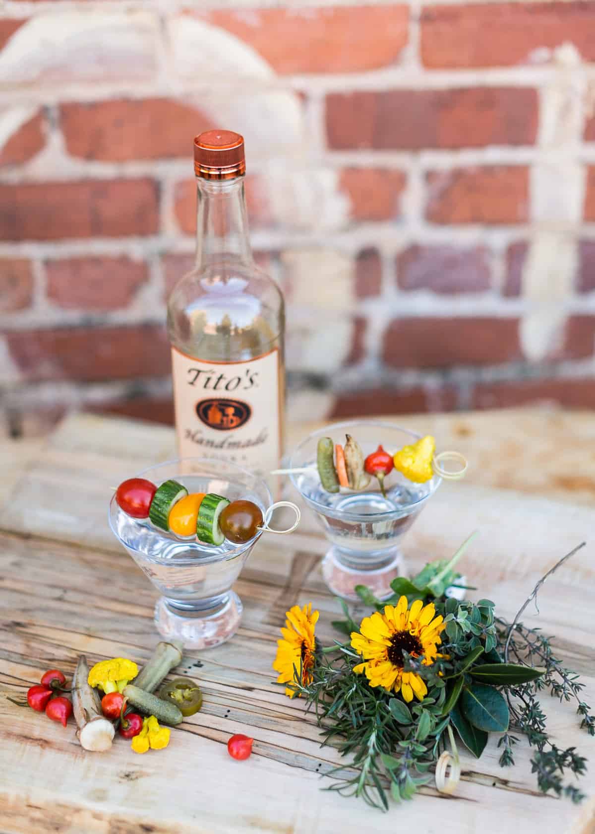 Tito's vodka bottle with two cocktails with fresh vegetables and other garnish on a wooden board.