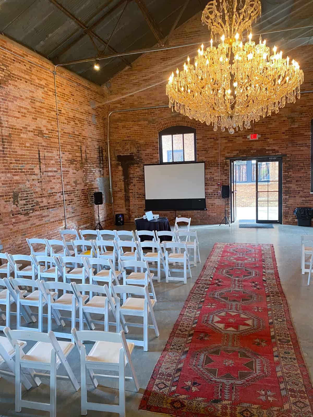 Empire Mills Marsden hall set up for an event with chairs, a screen, an oriental rug, and a large elegant chandelier.