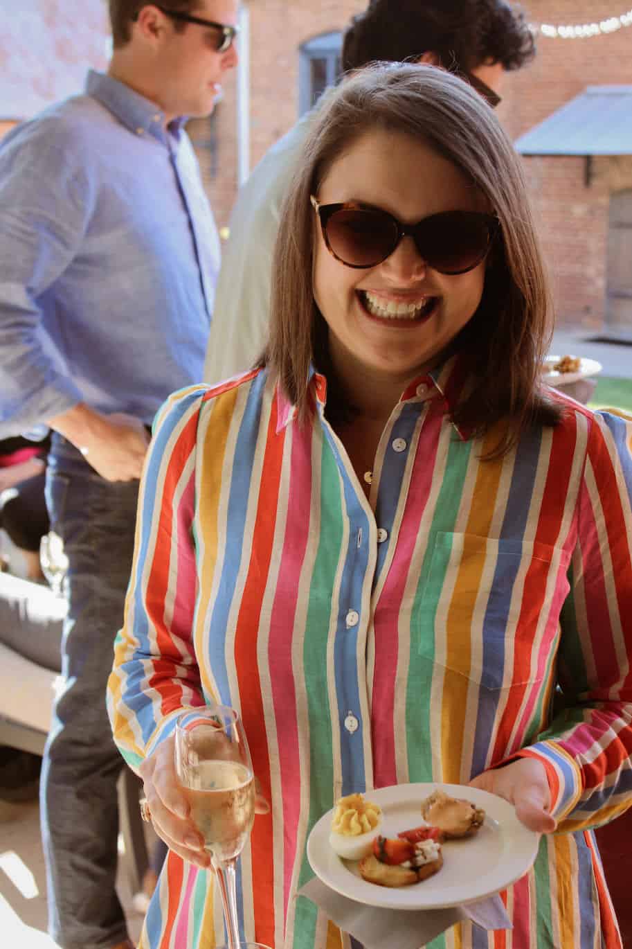 A woman in a colorful rainbow dress and sunglasses holding a glass of champagne and a plate of appetizers enjoying herself at an Empire Mills event